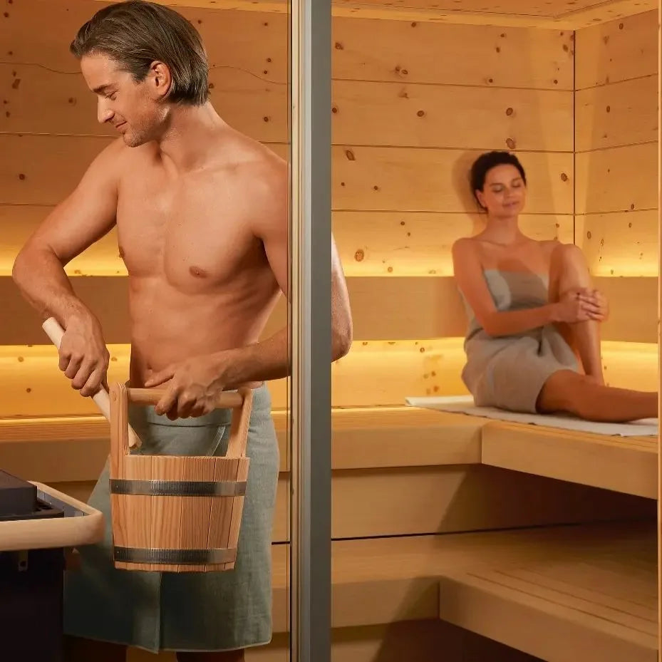 Man carrying a wooden bucket into a sauna with a woman sitting inside.