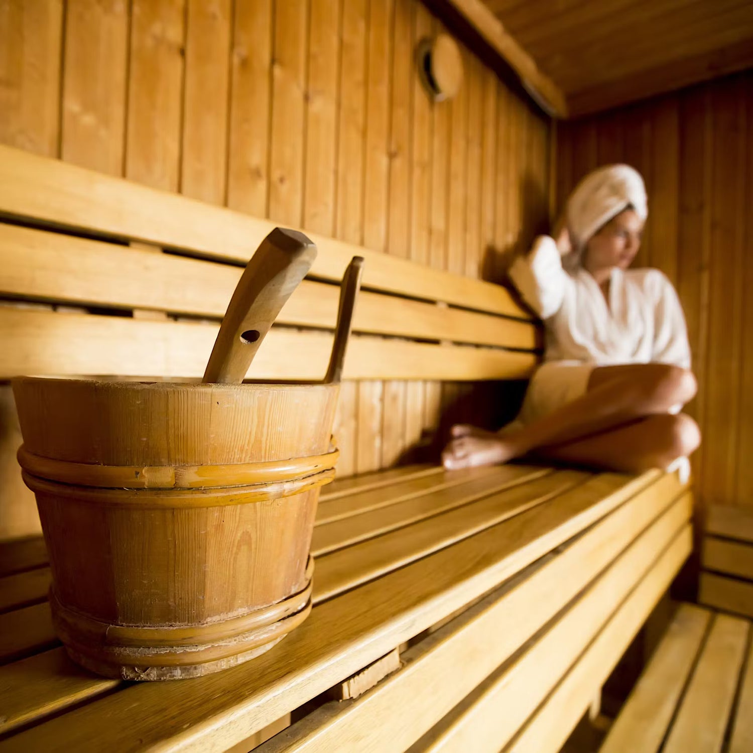 Person sitting on a wooden bench in a sauna with a wooden bucket and spoon nearby.