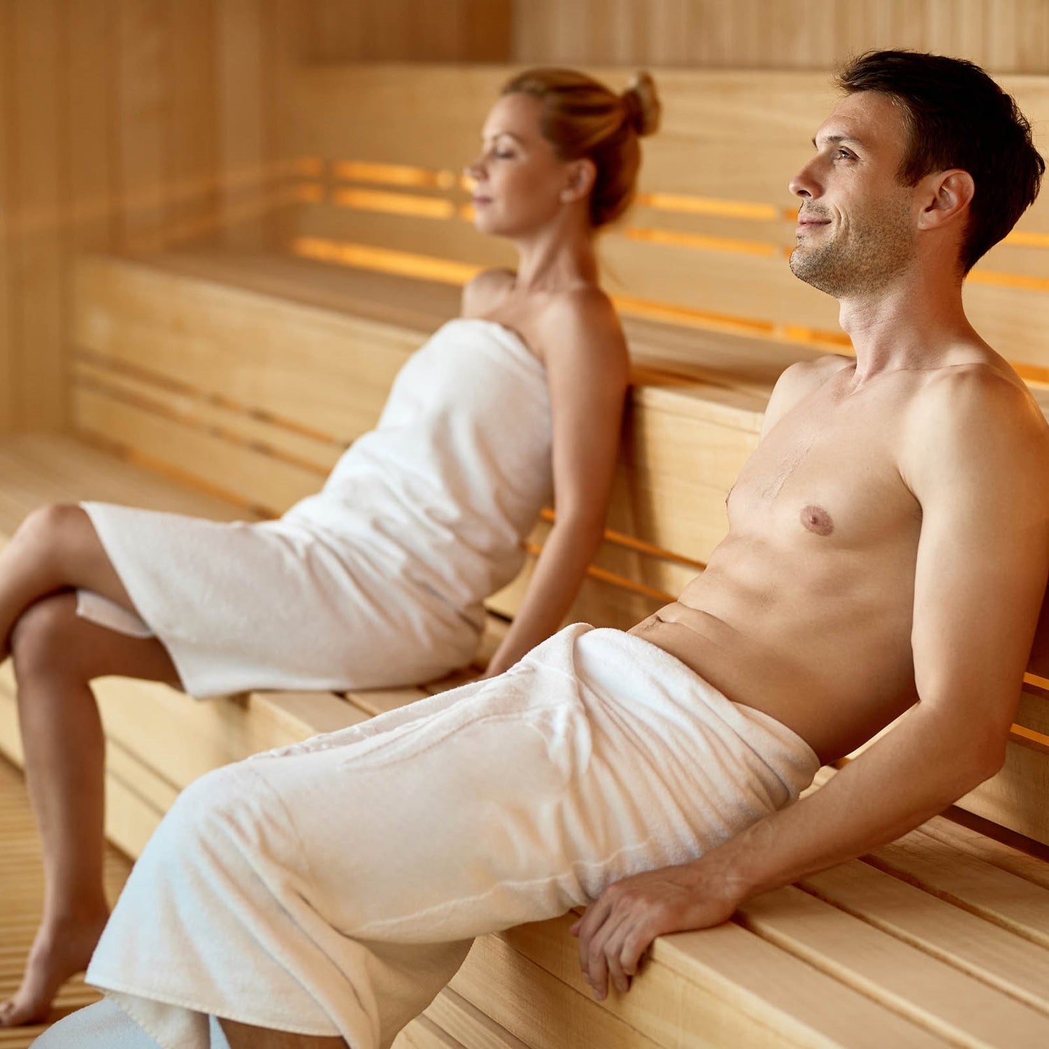 Man and woman sitting in a sauna with wooden benches and warm lighting.